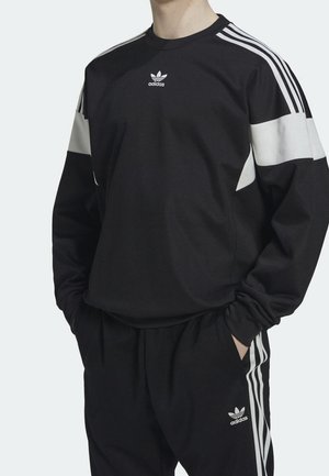 Person trägt schwarzen Adidas-Trainingsanzug mit weißen Streifen an Ärmeln und Hose, Hände in den Taschen, steht vor einem einfarbigen Hintergrund.
