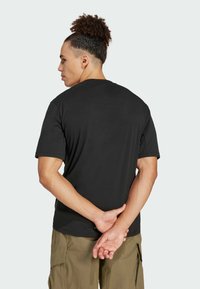 Man met krullend haar in een zwart T-shirt en kaki cargobroek, staand met handen samengevoegd achter zijn rug, wegkijkend.
