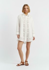 Abito camicia bianco con chiusura frontale a bottoni, ricami al centro e maniche lunghe. Abbinato a sandali bassi marroni.