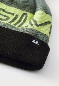 Un bonnet tricoté en vert et jaune avec un revers noir. Présente un motif en vagues et une étiquette de logo dans la section noire. Texture douce.