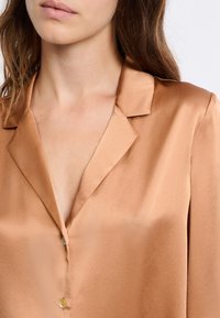 Satynowa, jasnobrązowa bluzka z kołnierzykiem z wycięciem i plisą zapinaną na guziki. Tkanina ma gładką fakturę i delikatny połysk.
