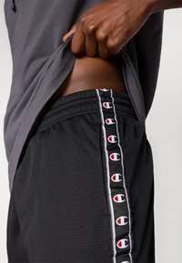 Μαύροι αθλητικοί σορτς από δίχτυ με κόκκινες και λευκές λογότυπες λεπτομέρειες στο πλάι, ελαστική μέση και ελαφρύ, αεριζόμενο ύφασμα.