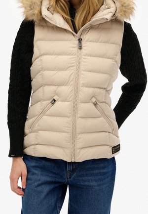 Woman wearing beige quilted sleeveless vest with fur-trimmed hood, black cable-knit sweater, and blue jeans, standing with left hand by side.