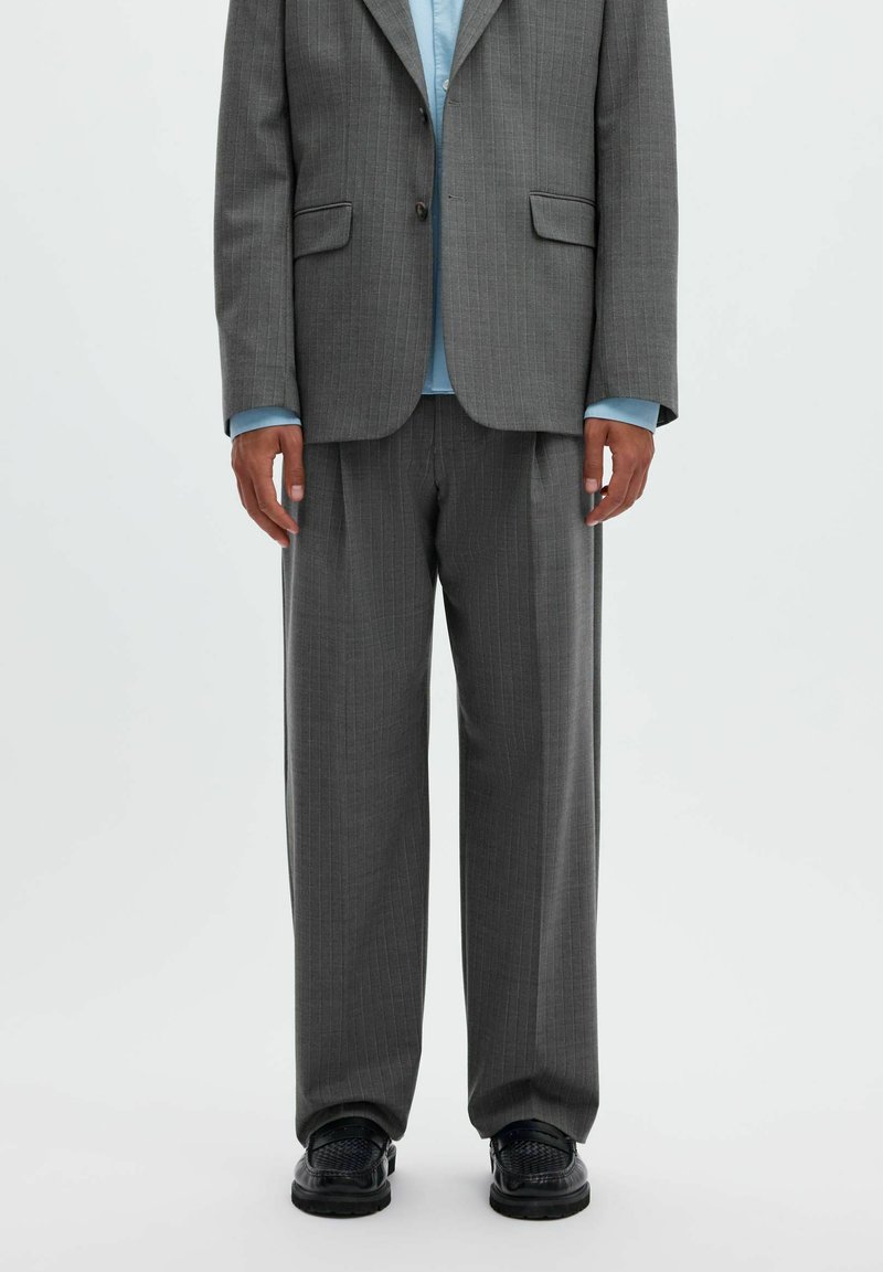 Homme portant un costume gris à fines rayures avec un pantalon ample, une chemise bleu clair et des chaussures en cuir tissé noires, debout devant un fond blanc.