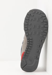 Semelle en caoutchouc grise avec des profondeurs de crantage, détail de talon courbé et accents rouges. Tige en daim avec des détails bleu marine sur le côté, conçue pour l'adhérence.