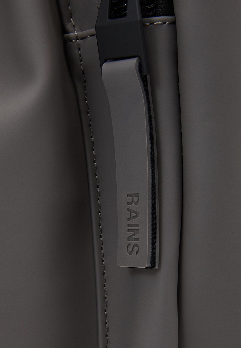 Dettaglio ravvicinato di una borsa waterproof grigia, con una texture liscia, etichetta in rilievo "RAINS" e inserti in hardware nero lungo le cuciture.