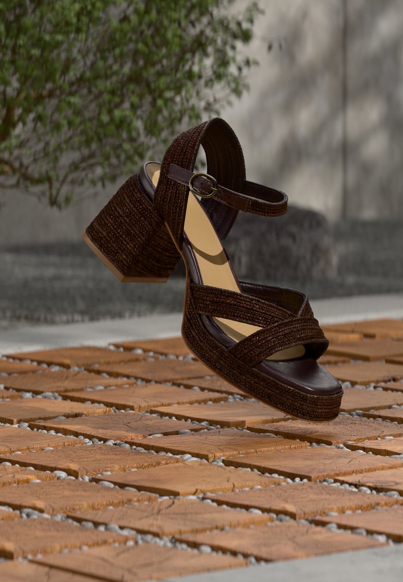 Καφέ πλεκτό σανδάλι με τακούνι μπλοκ και λουράκι στον αστράγαλο, αιωρούμενο πάνω από ξύλινη πλακόστρωτη επιφάνεια σε εξωτερικό χώρο, με θολό πράσινο φόντο στο βάθος.