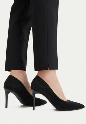 Kobieta nosząca czarne zamszowe szpilki z ostrym noskiem, zestawione z czarnymi, krótkimi spodniami z małym rozcięciem po bokach przy dole nogawek.