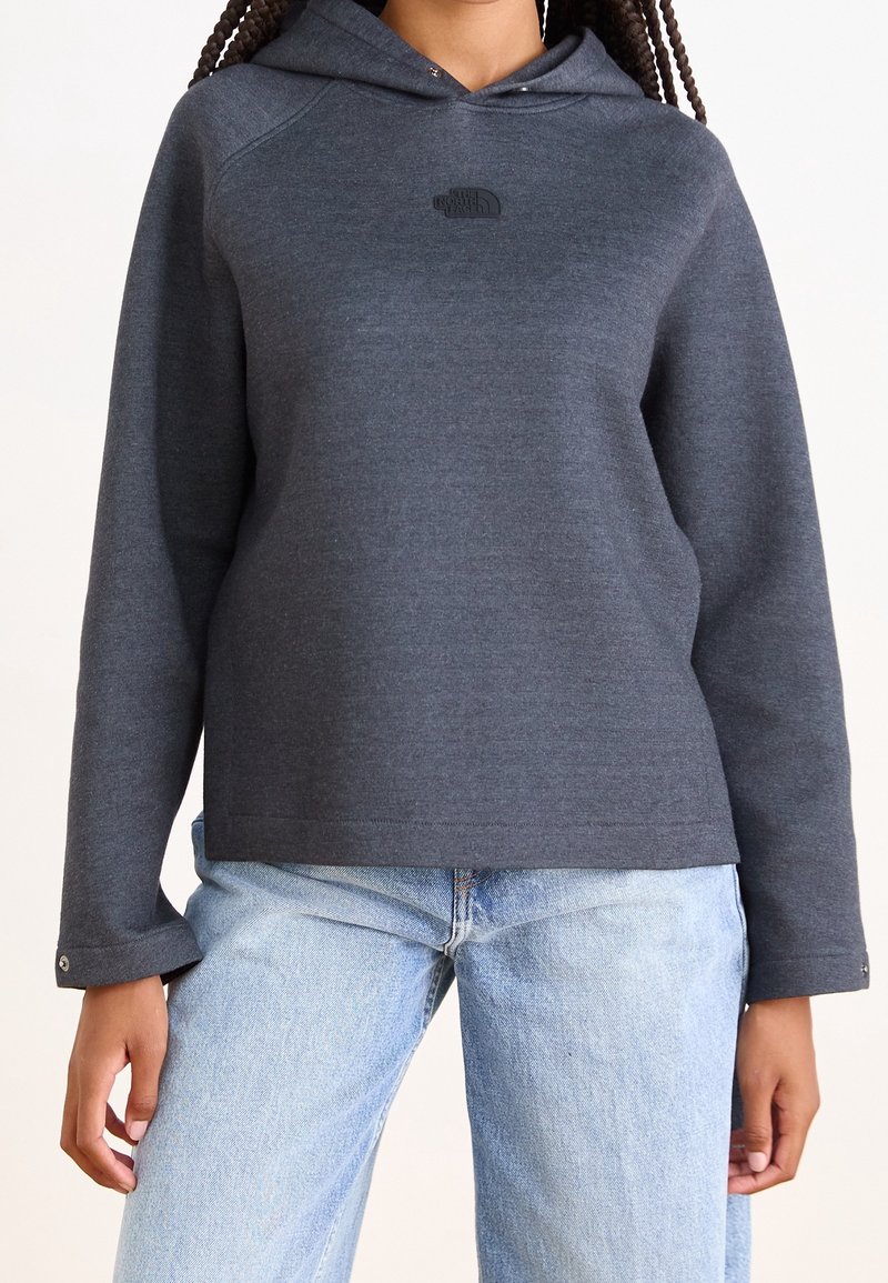 Personne portant un sweat à capuche gris foncé avec un petit logo sur la poitrine et un jean bleu clair, debout devant un fond uni.