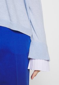 Maglione azzurro chiaro a maglia con polsini larghi, indossato sopra una blusa bianca a pieghe. Sullo sfondo si vede un tessuto blu navy liscio.