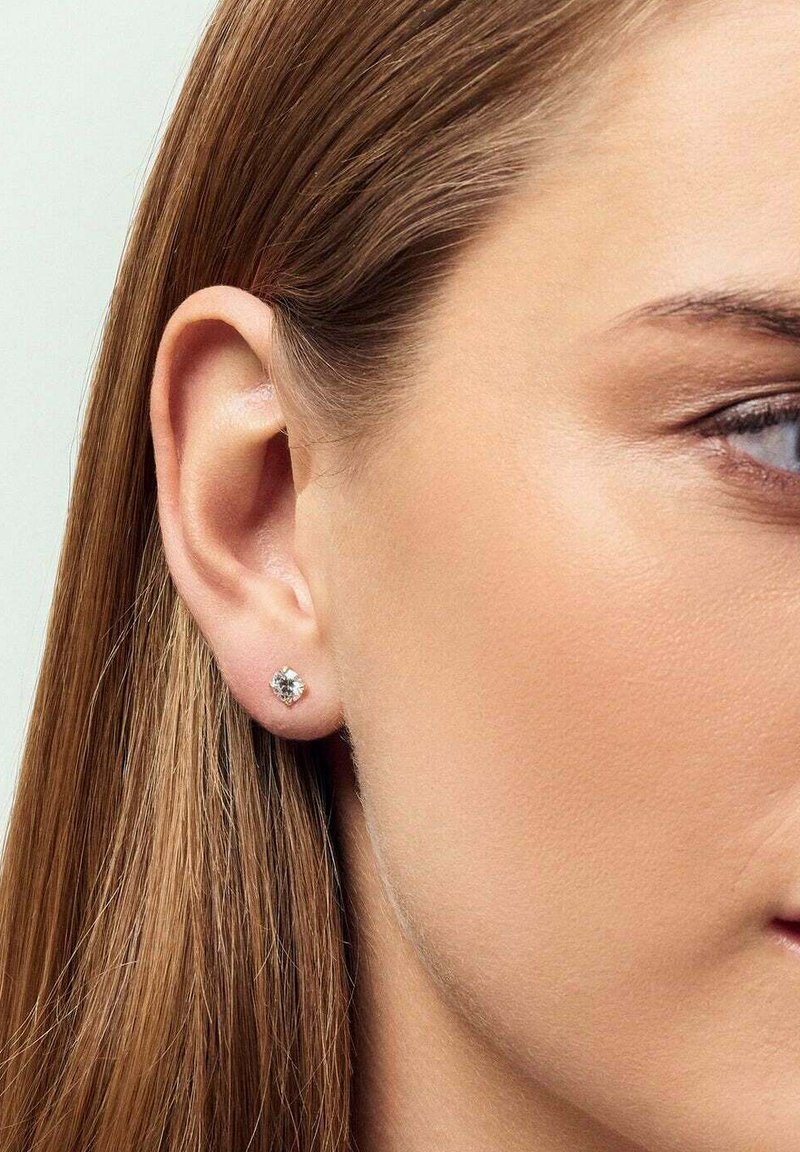 Silver stud earrings med en klar ädelsten, i en fyrkantig design, bärs i örat. Slätt hår faller ner bredvid örat.