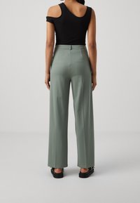Czarna bluzka na ramiączkach i szerokie, zielone spodnie o gładkiej fakturze. Spodnie mają wysoki stan i są zestawione z czarnymi butami.