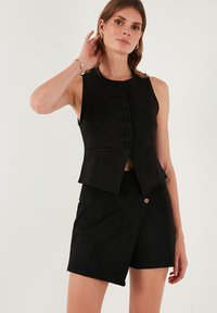 Vesta neagră fără mâneci, cu nasturi în față și buzunare, asociată cu pantaloni scurți negri cu talie înaltă, având un sistem de închidere cu un singur nasture. Textură din suede.