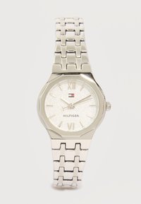 Ασημένιο μεταλλικό ρολόι χειρός με συνδεδεμένο μπρασελέ, υφασμένο στρογγυλό καντράν, ρωμαϊκοί αριθμοί, τρία δείκτες και μικρό λογότυπο Tommy Hilfiger κάτω από τις 12 το μεσημέρι.