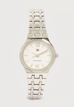 Ασημένιο μεταλλικό ρολόι χειρός με συνδεδεμένο μπρασελέ, υφασμένο στρογγυλό καντράν, ρωμαϊκοί αριθμοί, τρία δείκτες και μικρό λογότυπο Tommy Hilfiger κάτω από τις 12 το μεσημέρι.