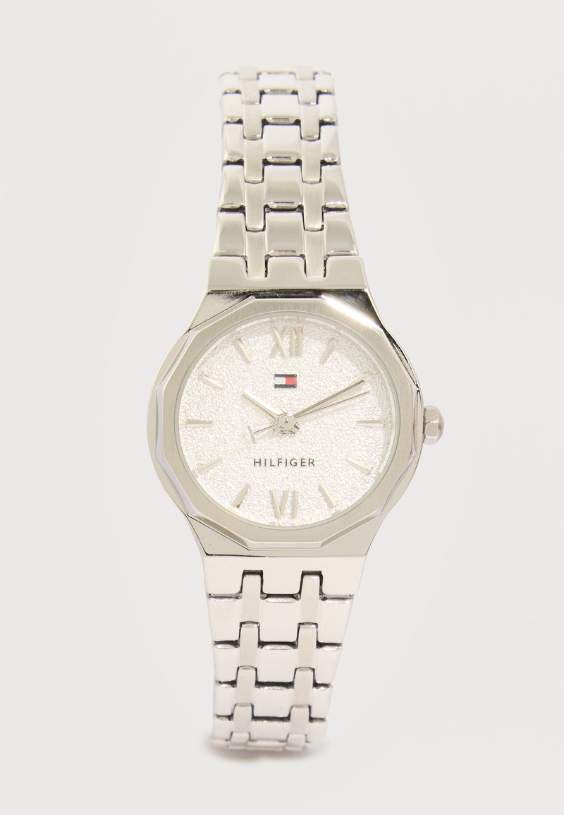 Ασημένιο μεταλλικό ρολόι χειρός με συνδεδεμένο μπρασελέ, υφασμένο στρογγυλό καντράν, ρωμαϊκοί αριθμοί, τρία δείκτες και μικρό λογότυπο Tommy Hilfiger κάτω από τις 12 το μεσημέρι.