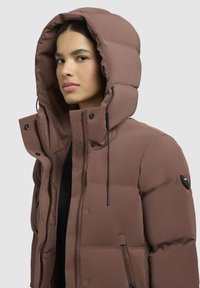 Pruun puffer-jakk suure kapuutsiga, külje-taskutega ja lukuga sulgemisega. Sile tekstuur peente õmbluste ja logo detailiga küljel.
