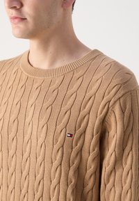 Gros plan d'une personne portant un pull beige torsadé avec un petit logo rectangulaire rouge, blanc et bleu sur la poitrine.