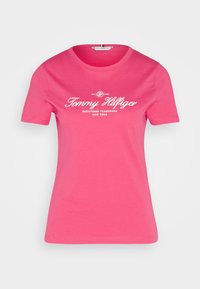 Rosa bomulds T-shirt med korte ærmer og rund halsudskæring. Har et hvidt tekst logo "Tommy Hilfiger" i midten, i en vintage stil.
