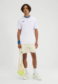 Weißes T-Shirt mit blauer Kragen und Logo, beigefarbene Shorts, weiße Sneakers und eine Netztasche, die einen leuchtend grünen Ball hält. Lockiges Haar.
