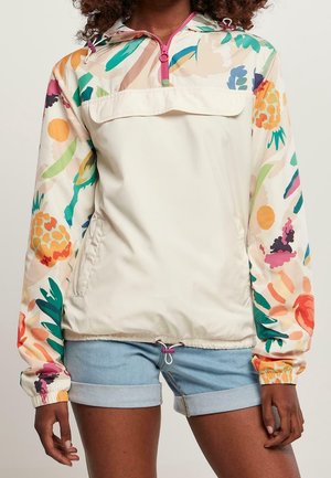 Femme portant un pull écru avec des manches imprimées de fleurs et de fruits colorés et un short en jean bleu retroussé.