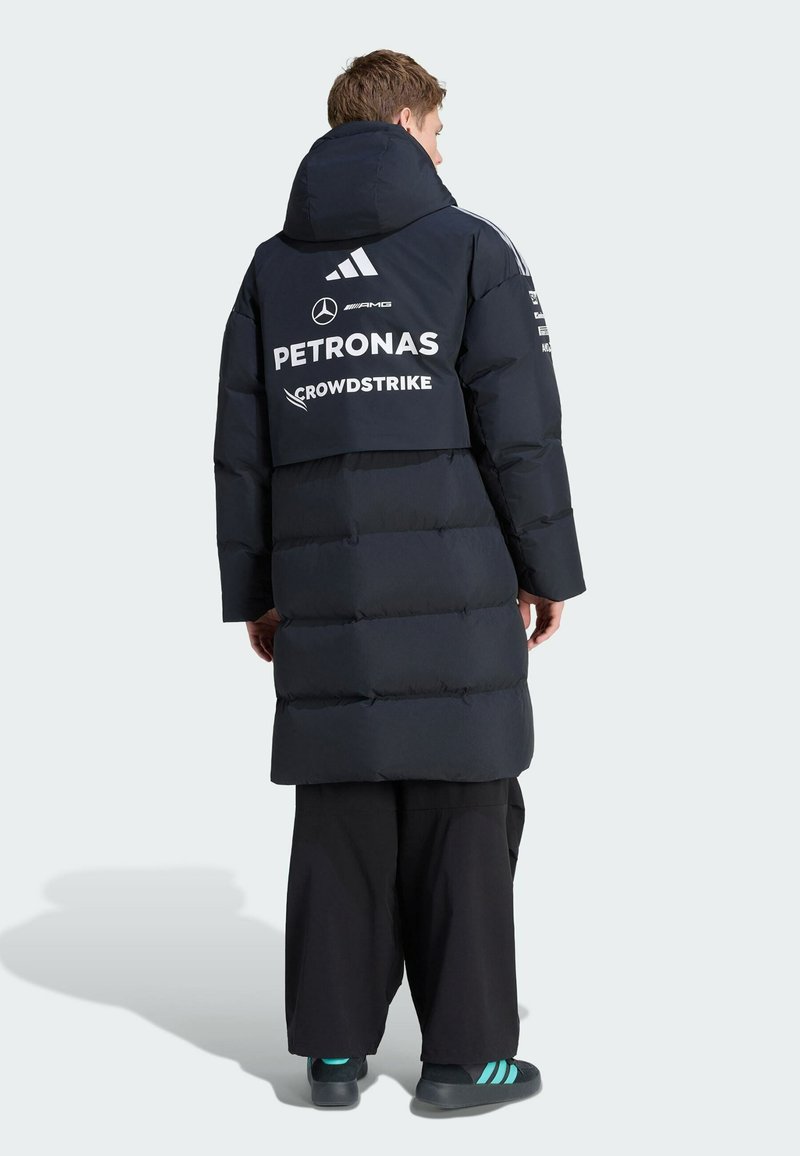 Persoon draagt een lange zwarte gewatteerde jas met capuchon, met Mercedes AMG Petronas en Crowdstrike-logo's op de achterkant, gecombineerd met een zwarte broek en sneakers.