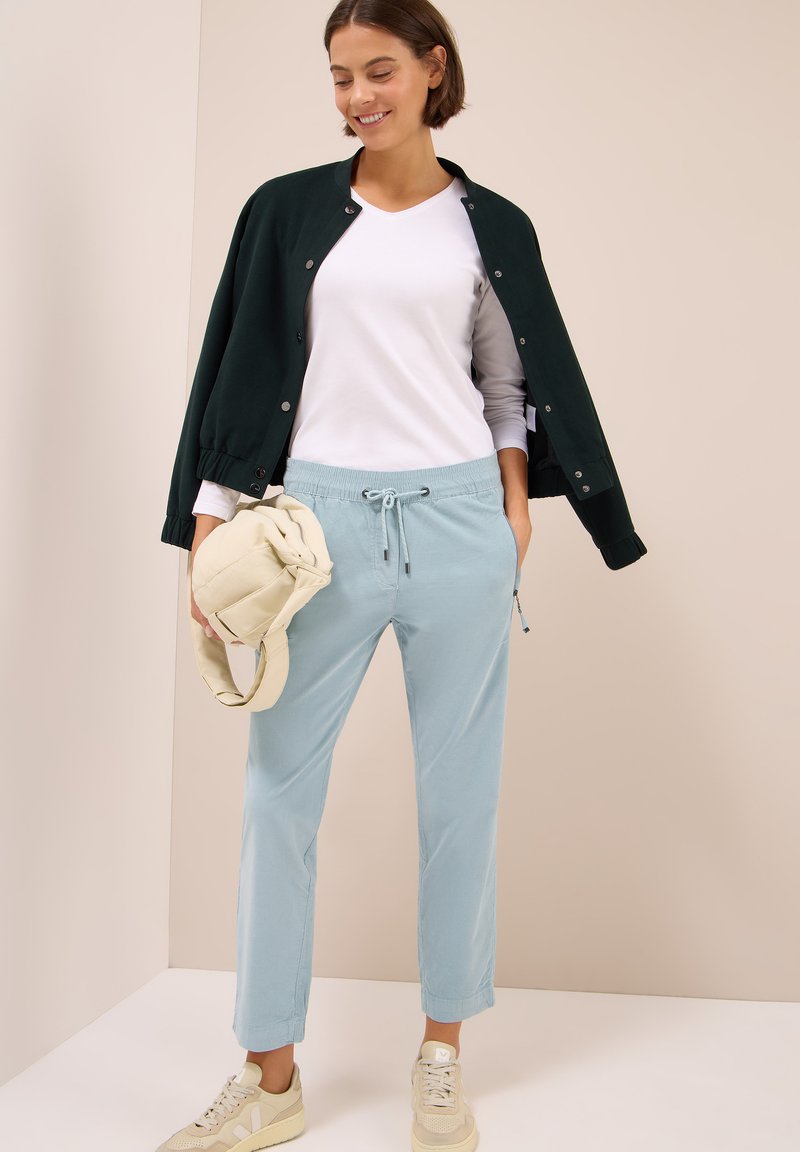 Pantalons de jogging bleu clair avec taille à cordon, t-shirt blanc en col V, et veste bomber verte foncée, associés à des baskets beige.