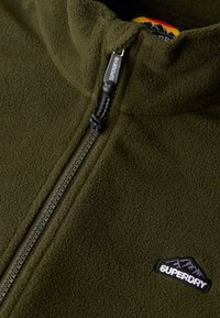 Zielona polarowa kurtka z wysokim kołnierzem, zamkiem błyskawicznym i czarnym logo górskim. Miękka faktura i szczegółowe przeszycia wzdłuż zamka.