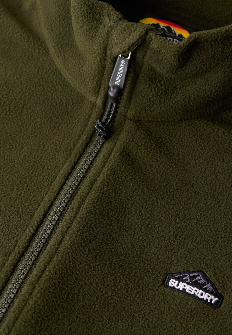 Veste en polaire verte avec un col haut, une fermeture éclair et un logo de montagne noir. Texture douce et couture détaillée le long de la fermeture éclair.
