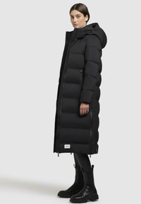Abrigo acolchado negro con un diseño largo y estructurado, patrón de acolchado vertical, capucha y bolsillos laterales. Lleva puesto sobre leggings negros y botas.