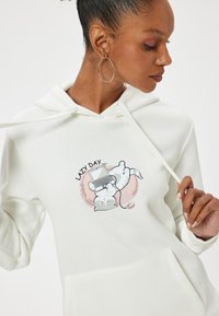 Sudadera blanca con un gráfico de un gato con una laptop y el texto "DÍA DE PEREZA...". El material parece suave, con cordones y un bolsillo frontal.