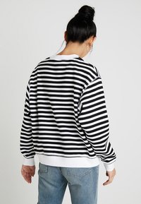 Kvinna med svart hår i en knut, klädd i en svart och vit horisontellt randig tröja och ljusblå jeans, vänd bort mot en enfärgad bakgrund.