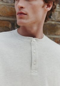 Valkoisen ja kellertävän sävyinen waffle-neulottu henley-paita, jossa on pyöreä pääntie ja kolmen napin läppä, tekstuuripintainen kangas.