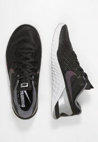 Mustat Nike-kuntosalitossut, joissa on teksturoidut pinnat, valkoiset pohjat ja heijastavat yksityiskohdat. Sisältää nauhakiristysjärjestelmän ja pehmustetun varren.