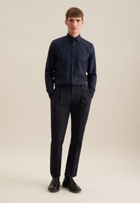 Camicia da uomo blu scuro con colletto button-down, abbinata a pantaloni neri su misura e scarpe stringate nere, su uno sfondo neutro.