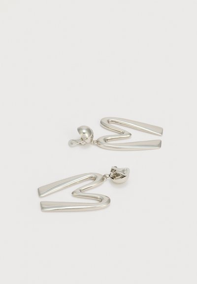 Ασημένια μεταλλικά σκουλαρίκια με κλιπ, με αφηρημένα, γωνιώδη ζιγκ-ζαγκ σχέδια σε απλό λευκό φόντο.