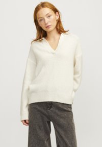 Maglione bianco, morbido e dal taglio ampio con scollatura a V e maniche lunghe, realizzato in tessuto testurizzato. Indossato sopra pantaloni grigi a vita alta.