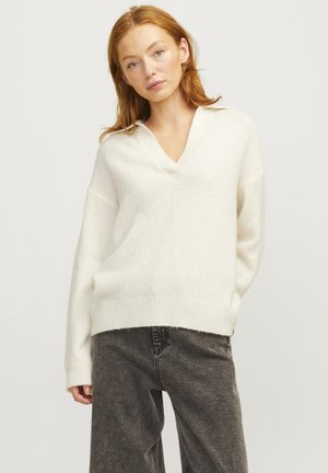 Pull blanc ample avec un col en V et des manches longues, fabriqué en tissu doux et texturé. Porté sur un pantalon gris taille haute.