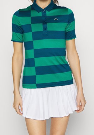 Grün und marineblau gestreiftes Poloshirt mit Kragen und drei Knöpfen, kombiniert mit einem weißen Faltenrock. Lacoste-Logo auf der linken Brust.