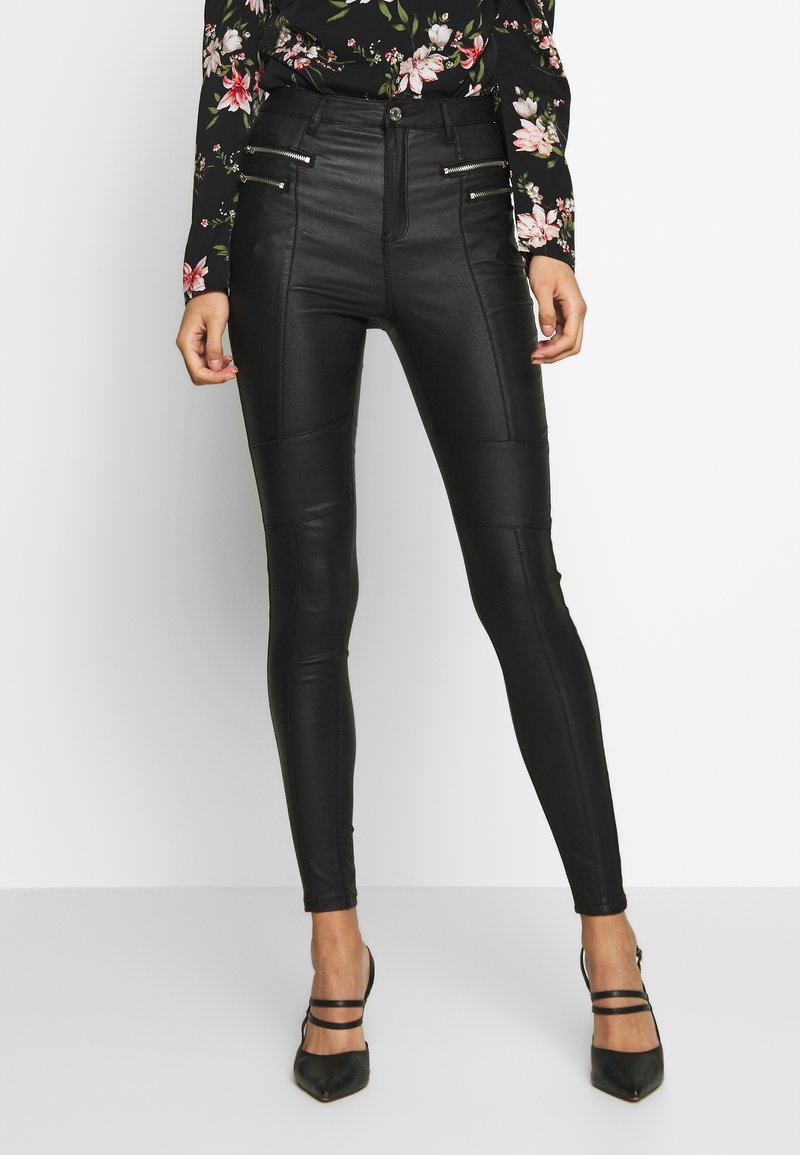 Pantalons skinny en simili cuir noir avec une texture lisse, dotés d'accents zippés et d'une taille haute, associés à des escarpins noirs à bouts pointus.