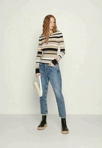 Top de mangas longas às riscas em bege, preto e creme; design ajustado. Jeans azuis de cintura alta com bainha desfiada. Botas ankle pretas e um chapéu.