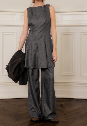 Vrouw draagt een grijs mouwloos tuniek en bijpassende broek met wijd uitlopende pijpen, houdt een zwart jasje vast, staat op een houten vloer bij een witte geprofileerde muur.