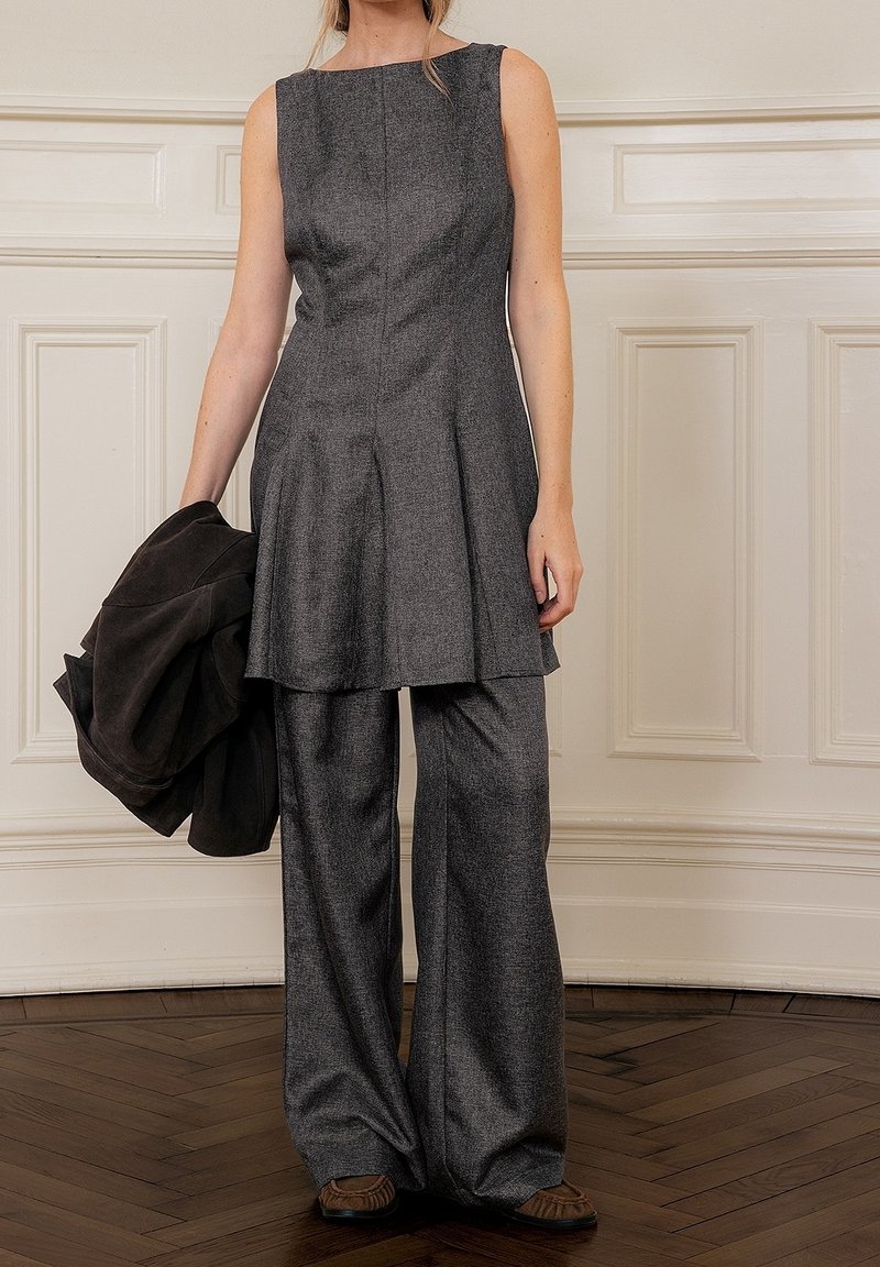 Mujer con túnica gris sin mangas y pantalones anchos a juego, sosteniendo una chaqueta negra, de pie sobre suelo de madera junto a una pared blanca con paneles.