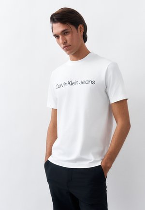 Man met halflang donker haar draagt een wit Calvin Klein Jeans T-shirt en een zwarte broek, staand met handen in de zakken tegen een witte achtergrond.