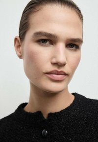 Zbliżenie osoby w teksturowanym czarnym ubraniu z okrągłym dekoltem i pojedynczym zapięciem na guzik, włosy zaczesane do tyłu, makijaż w neutralnych odcieniach.