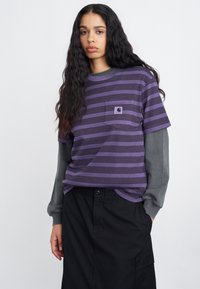 Vijolična in črna prosta majica s kratkimi rokavi s žepom na prsih, nošena preko sivega dolgoročnega topa. Tkanina je mehka bombažna z udobnim krojem.