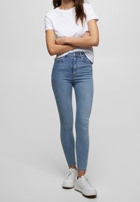 Jeans ajustados de mezclilla azul con cintura a media altura, combinados con una camiseta blanca y zapatillas blancas, que muestran una silueta ceñida y un ligero estiramiento.