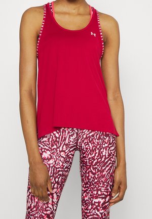Femme portant un haut sans manches rouge Under Armour et des leggings roses à motifs, montrée du cou jusqu'au milieu de la cuisse sur fond uni.