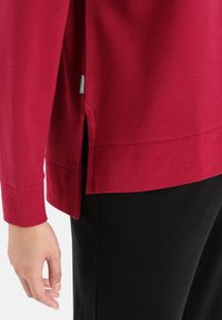 Suéter rojo de lana con una abertura lateral y puños acanalados, que presenta una etiqueta visible. Llevado sobre pantalones negros, mostrando un ajuste relajado.