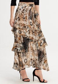 Jupe maxi en tissu transparent à motifs avec des volants superposés. Les couleurs incluent le marron, le beige et le noir avec des motifs floraux et abstraits.